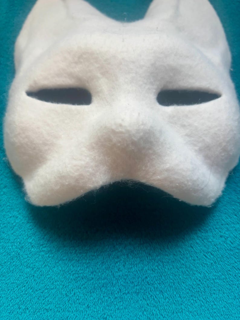 May include: A white felt mask featuring a stylized animal shape with pointed ears and two horizontal, almond-shaped eye openings. The surface has a soft, fuzzy texture, and the mask is molded to follow the contours of a face with a defined nose bridge.