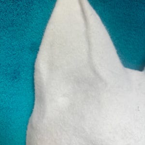 May include: A white felt mask featuring pointed, upright ears and a smooth, soft texture. The mask has a distinct, angular shape with a cutout section visible at the bottom, showcasing a clean, minimalist design crafted from dense, white fabric material.