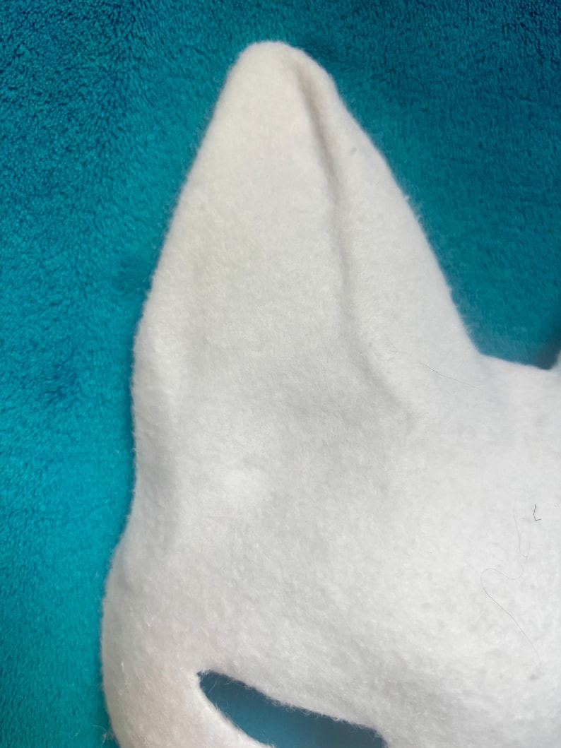 May include: A white felt mask featuring pointed, upright ears and a smooth, soft texture. The mask has a distinct, angular shape with a cutout section visible at the bottom, showcasing a clean, minimalist design crafted from dense, white fabric material.