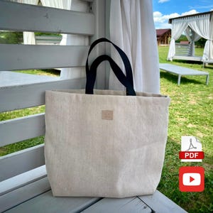 Könnte beinhalten: Beige Tragetasche mit schwarzen Henkeln, auf einer Holzoberfläche. Die Tasche hat einen kleinen, quadratischen Aufnäher unten. Im Hintergrund ein weißer Vorhang und ein Pavillon.
