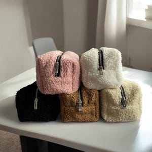 Puede incluir: Cinco bolsas de cremallera peludas en diferentes colores: negro, marrón, crema, blanco y rosa. Las bolsas están apiladas unas encima de otras.