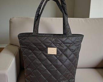 May include: A black quilted tote bag with two handles. The bag features a diamond pattern and a small rectangular patch with text. The bag is on a beige sofa.
