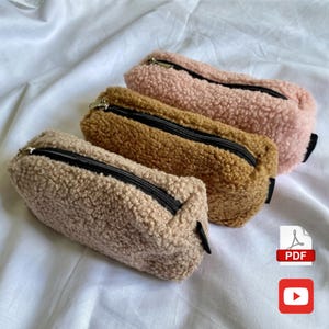 May include: Three fluffy pencil cases in shades of beige, brown, and pink. Each case has a black zipper and a small black tab on one end. The cases are made of a soft, textured material.