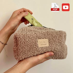 Può includere: Un beauty case rettangolare beige e soffice con chiusura a cerniera. La borsa ha una toppa in pelle con la scritta "VD&O Made in Ukraine". Una mano sta inserendo un oggetto verde nella borsa. Un'icona PDF e un'icona di YouTube si trovano nell'angolo in alto a destra.