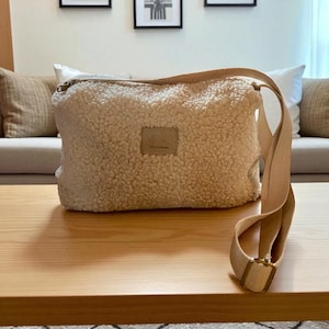 Puede incluir: Un bolso bandolera beige y esponjoso con una correa larga y ajustable y una hebilla rectangular. El bolso tiene un pequeño parche cuadrado con texto en la parte delantera. El bolso está sobre una mesa de madera.