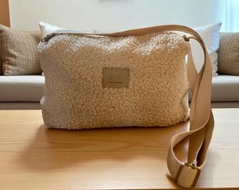 May include: A beige, fluffy crossbody bag with a long, adjustable strap and a rectangular buckle. The bag has a small, square patch with text on the front. The bag is sitting on a wooden table.