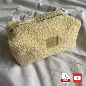 May include: A rectangular, cream-colored, fluffy cosmetic bag with a black zipper. The bag has a textured, sherpa-like exterior and a small, rectangular label with the text "V&O" and "Made in Ukraine".