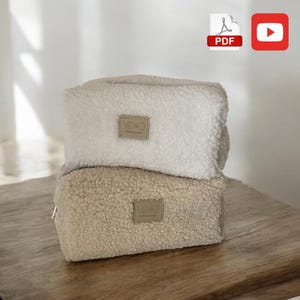 Può includere: Due morbide trousse rettangolari impilate su una superficie di legno. La borsa superiore è bianco sporco e quella inferiore è beige. Ogni borsa ha una piccola etichetta rettangolare con la scritta "VD&O".