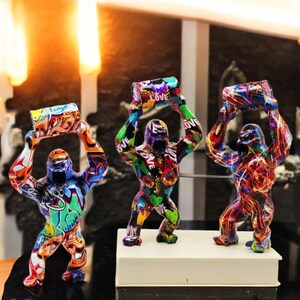 Peut inclure: Trois sculptures de gorilles colorées de style graffiti, chacune tenant un objet cylindrique au-dessus de sa tête. Les sculptures sont en résine et ont une finition brillante. Les objets qu'ils tiennent sont également recouverts de motifs de style graffiti.