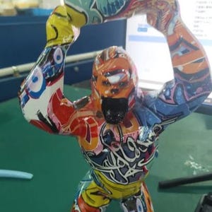 May include: A gorilla figurine standing upright with both arms raised above its head, covered in a vibrant, multi-colored graffiti pattern featuring shades of blue, orange, yellow, red, and white with black outlines and stylized lettering.