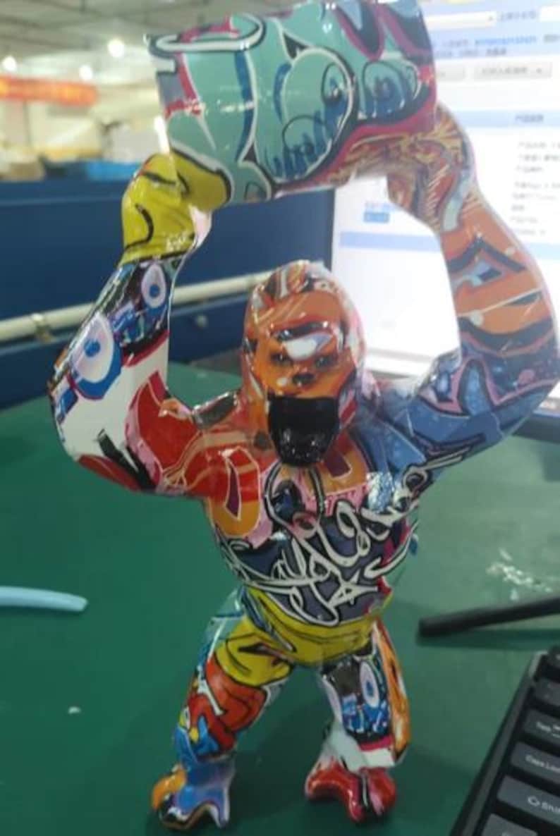 May include: A gorilla figurine standing upright with both arms raised above its head, covered in a vibrant, multi-colored graffiti pattern featuring shades of blue, orange, yellow, red, and white with black outlines and stylized lettering.