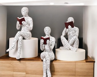 Könnte beinhalten: Drei weiße Keramikskulpturen von Menschen, die sitzen und Bücher lesen. Die Skulpturen sind mit schwarzem Text bedeckt und stehen auf weißen Sockeln. Die Skulpturen stehen auf einer Holzoberfläche.