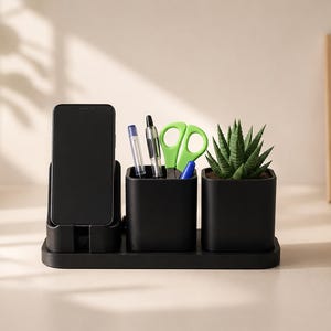 May include: A matte black desk organiser set featuring a vertical smartphone stand, a square pen holder containing pens and green scissors, and a square planter holding a small green succulent plant, all mounted on a rectangular base.