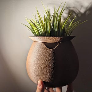 May include: A brown, textured ceramic pot with a wide rim, holding vibrant green and yellow artificial grass. The pot has a unique shape with a curved body and a small opening. The pot is held by a hand.