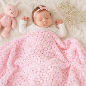 May include: A sleeping baby wearing a pink bow headband and a white onesie is covered with a pink, open-weave blanket. A pink teddy bear and white flowers are also in the image, creating a soft, gentle scene.