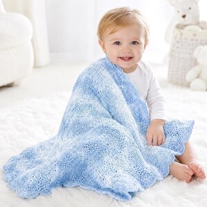 May include: A baby wrapped in a light blue and white crocheted blanket with a scalloped edge. The blanket has a delicate, open weave pattern. The baby is wearing a white onesie and is sitting on a white, fluffy rug.