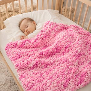 May include: A chunky knit baby blanket featuring a variegated pattern of soft pink, bright pink, and white yarn. The blanket has a thick, textured loop stitch construction and is draped over a sleeping infant resting on a white pillow in a crib.