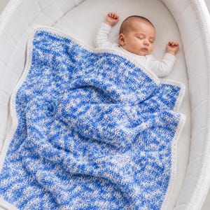 May include: A baby blanket featuring a variegated pattern of blue and white yarn with a solid white crocheted border. The blanket is draped over a sleeping infant who is wearing a long-sleeved white shirt while lying in a white bassinet.