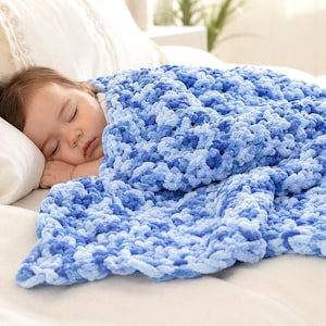 Op de afbeelding: Een slapende baby onder een zachte, blauw-witte gehaakte deken. De deken heeft een getextureerd, pluche uiterlijk, met verschillende tinten blauw. De baby rust op een wit kussen en laken.