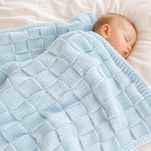 May include: A baby sleeps peacefully under a light blue knitted blanket. The blanket has a textured, square pattern. The soft blanket is draped over the baby, creating a cozy and comforting scene.
