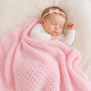 May include: A sleeping baby wrapped in a soft, light pink knitted blanket. The baby wears a white long-sleeved onesie and a pink headband with a bow. The blanket has a delicate openwork pattern.