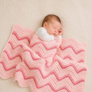 Peut inclure: Un bébé endormi enveloppé dans une couverture en crochet rose et rose clair à motif chevron. La couverture a un bord festonné. Le bébé porte un haut blanc à manches longues.