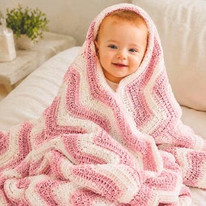 May include: A baby wrapped in a handmade crochet blanket featuring a repeating chevron pattern in alternating stripes of soft pink and cream white yarn. The blanket covers the head and shoulders, showcasing a textured stitch design throughout.