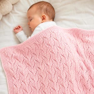May include: A sleeping baby is covered with a soft, pink knitted blanket. The blanket features a repeating diamond pattern with openwork details. The baby is wearing a white onesie and is resting on a white sheet.