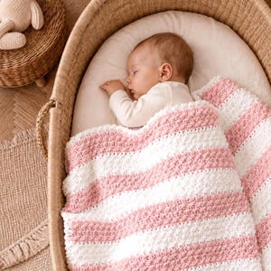 May include: A sleeping baby is nestled in a wicker bassinet, covered with a pink and white striped blanket. The baby is wearing a white onesie. A woven basket and a small rug are visible in the background.