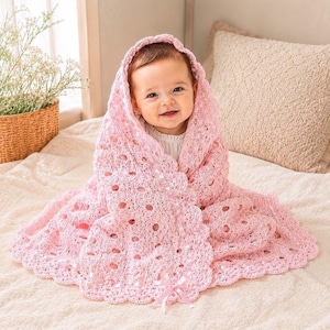 May include: A light pink crochet baby blanket with a delicate openwork pattern and a scalloped edge. The blanket features a woven satin ribbon detail along the border, tied into a small bow at the bottom center, providing a soft texture.