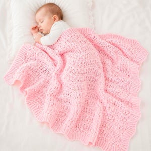May include: A sleeping baby is nestled under a soft, pink, knitted blanket. The blanket has a delicate openwork design and a scalloped edge. The baby is wearing a white onesie and resting on a white pillow.