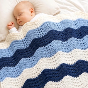 May include: A crochet baby blanket featuring a wavy ripple pattern in alternating stripes of cream, light blue, and dark navy blue yarn. The blanket has a textured, open-stitch design and is draped over a sleeping infant on a white pillow.