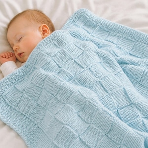 Può includere: Un bambino che dorme è rannicchiato sotto una coperta di maglia azzurra. La coperta presenta un motivo strutturato e intrecciato e un bordo spesso. Il bambino indossa un body bianco e riposa su un lenzuolo bianco.