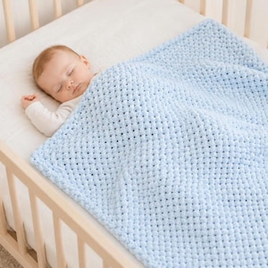 Op de afbeelding: Een slapende baby ligt in een wieg, bedekt met een zachte, lichtblauwe gebreide deken. De deken heeft een textuur, geweven patroon. De wieg is gemaakt van lichtgekleurd hout.