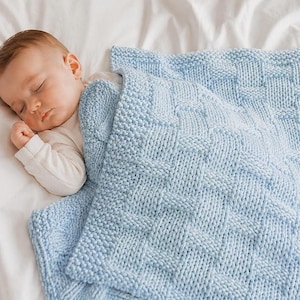 May include: A light blue baby blanket featuring a textured basketweave knit pattern. The blanket is crafted from thick yarn and displays a consistent square grid design throughout the fabric, providing a soft and cozy appearance for a sleeping infant.