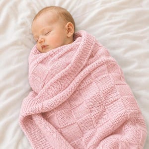 Puede incluir: Un bebé dormido envuelto en una suave manta de punto rosa claro. La manta tiene un patrón cuadrado texturizado. El bebé está acostado sobre una sábana blanca.