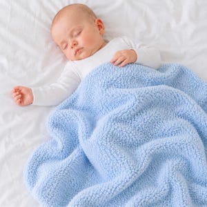 May include: A sleeping baby wearing a white long-sleeved onesie, covered with a soft, light blue blanket. The baby is lying on a white sheet. The blanket has a textured, plush appearance.