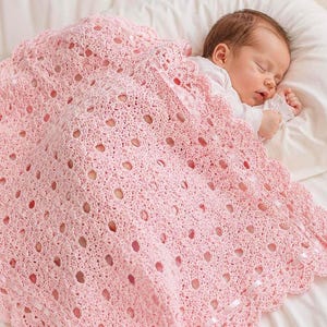 Può includere: Una coperta per neonati all'uncinetto rosa chiaro caratterizzata da un intricato motivo traforato con bordo smerlato e un dettaglio in nastro di raso intrecciato. La coperta è drappeggiata su un neonato che dorme con un capo bianco.