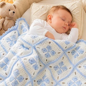 May include: A sleeping baby is nestled under a white and blue crocheted blanket with a floral pattern. A beige teddy bear with a bow is visible on the left. The baby is wearing a white long-sleeved shirt.