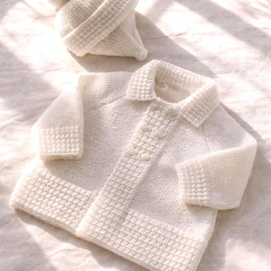 Peut inclure: Un ensemble pour bébé en tricot crème, comprenant un cardigan et un bonnet assorti. Le cardigan a un col et une fermeture à boutons. Le bonnet est orné d'un pompon. L'ensemble est en fil doux et texturé.