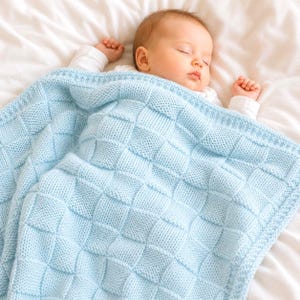 以下が含まれることがあります： 明るい青色のニットブランケットの下で眠っている赤ちゃん。ブランケットは、正方形の模様で織り込まれており、柔らかく快適に見えます。赤ちゃんは白いロンパースを着ています。