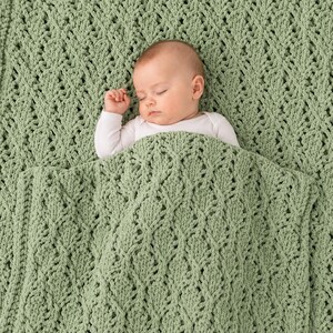 Puede incluir: Una manta de punto color verde salvia con un patrón de diamantes texturizado y un diseño de trenzas en relieve. La manta está hecha de hilo suave y proporciona una capa gruesa y acogedora. Un bebé descansa bajo la tela verde texturizada.