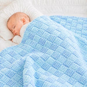 Könnte beinhalten: Ein schlafendes Baby ist mit einer blau-hellblauen Strickdecke bedeckt. Die Decke hat ein gewebtes Muster. Das Baby liegt auf einem weißen Kissen und trägt einen weißen Strampler.