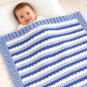 Pode incluir: Uma manta de bebé tricotada à mão com riscas horizontais em tons de azul escuro, azul claro e branco. A manta apresenta um padrão de pontos texturados em relevo no centro e uma borda canelada sólida em fio de cor azul claro.
