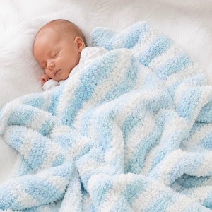 Puede incluir: Un bebé durmiendo envuelto en una manta suave y texturizada con rayas diagonales alternas de hilo azul claro y blanco. La manta tiene una apariencia afelpada y cubre al bebé, quien viste una camiseta blanca sencilla de manga larga.