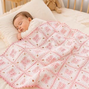 Può includere: Un bambino che dorme è rannicchiato sotto una coperta all'uncinetto bianca e rosa fatta a mano. La coperta presenta un motivo floreale ripetuto all'interno di blocchi quadrati. Il bordo della coperta è rifinito con un bordo smerlato, che aggiunge un tocco delicato al design morbido e accogliente.