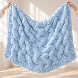 Puede incluir: Una manta tejida de color azul claro que presenta un patrón de trenzas gruesas con texturas verticales y un borde texturizado, confeccionada con hilo grueso en un tono azul pastel sólido y suave en toda la pieza.