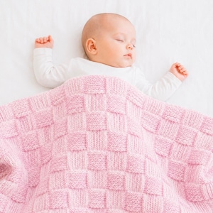 May include: A baby sleeps under a pink knitted blanket. The blanket features a woven square pattern. The baby wears a white long-sleeved top and rests on a white surface.