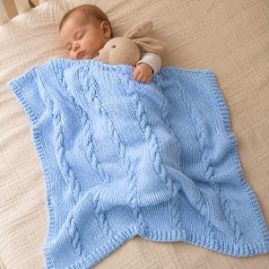 May include: A light blue knitted baby blanket featuring a vertical cable knit pattern and a ribbed border. The blanket is draped over a sleeping infant who is holding a small, tan-colored plush rabbit toy with long ears and a soft texture.