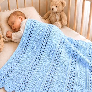 May include: A baby sleeps peacefully under a light blue, crocheted blanket in a crib. The blanket features a scalloped edge and vertical openwork stripes. A teddy bear sits in the crib, and a small basket is visible.
